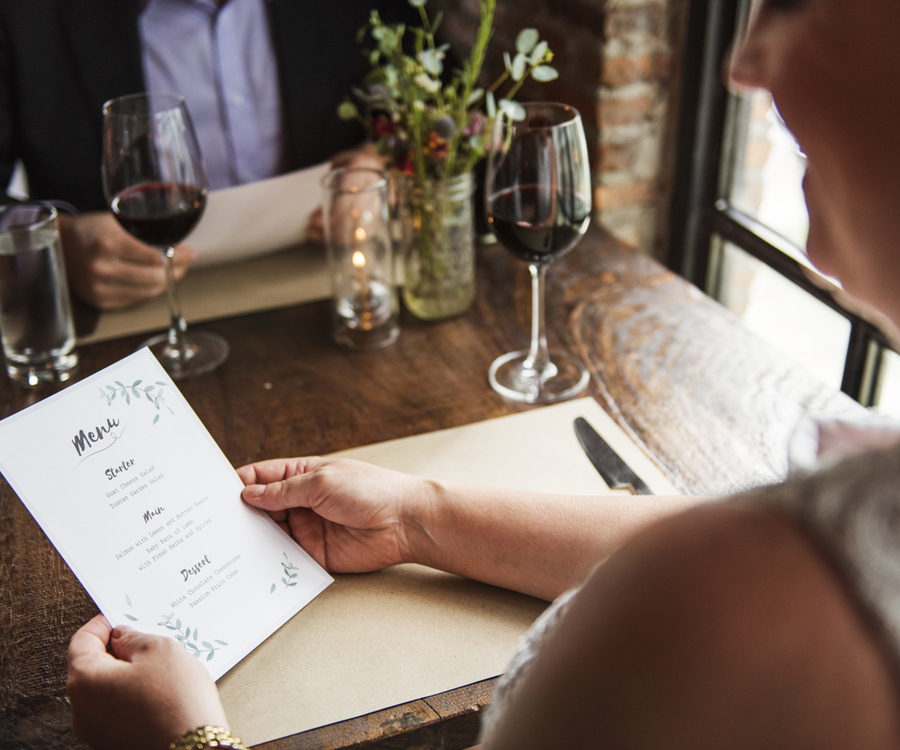 Woman reading a menu card with starter, main course and dessert at a wooden table in a restaurant, with a guest and a glass of red wine across from her