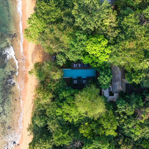 Aerial view of a secluded villa with a private pool, hidden in dense tropical forest beside a sandy beach and turquoise sea