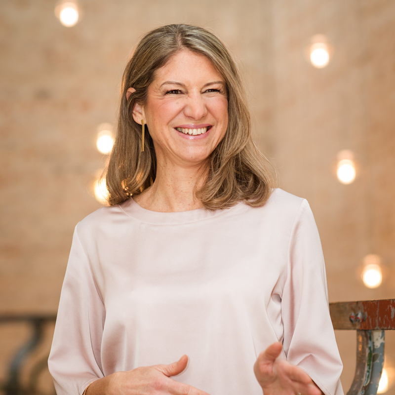 Portrait of TANIA SAEZ DE GUINOA, standing in a bright room with lights in the background and smiling